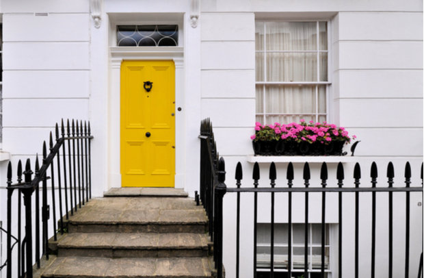 Close-up shot of a white house with a yellow front door and pink flowers on a window sill